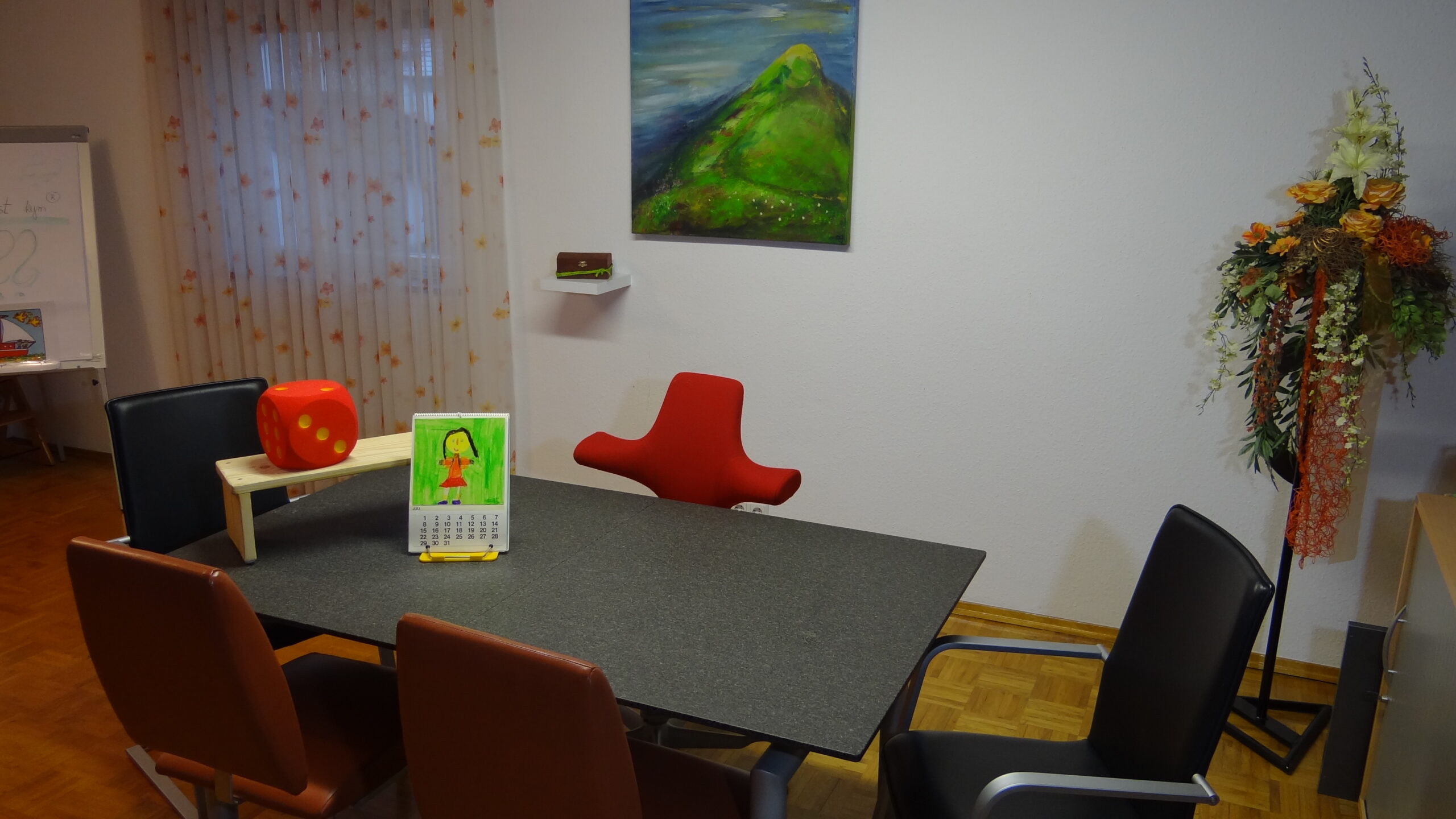 Freundlich eingerichteter Besprechungsraum mit Tisch, Stühlen, einem roten Würfel, einem Kinderkalender, einem Wandbild mit grüner Landschaft sowie einer großen Blumendekoration in der Ecke.